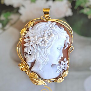 May include: A gold pendant with a white cameo depicting a woman's profile, decorated with flowers. The cameo is set on a brown background and framed by a gold floral design. The pendant includes a loop for a chain.
