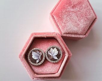 May include: A pair of round stud earrings with butterfly designs, set in a brown background and surrounded by a ring of small, clear stones. The earrings are displayed in an open, pink velvet hexagonal jewelry box, with a matching closed box in the background.