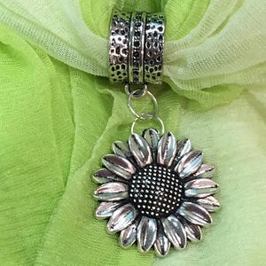 May include: A silver-toned scarf ring with a sunflower pendant. The ring has a textured design and connects to a detailed sunflower pendant. The background is a light green fabric.