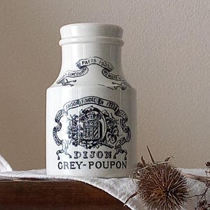 Pode incluir: Um pote de cerâmica branca com gargalo estreito e borda alargada. A frente apresenta um design impresso em preto com um brasão central, fitas e o texto "MAISON FONDEE EN 1777" e "DIJON GREY-POUPON" junto com "PARIS 1900" em uma faixa.
