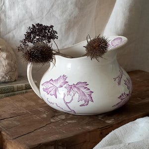 May include: A cream-coloured ceramic jug with a purple floral pattern, a handle, and a spout. The jug is on a wooden chopping board with dried flowers inside. An old fossil and books are in the background.