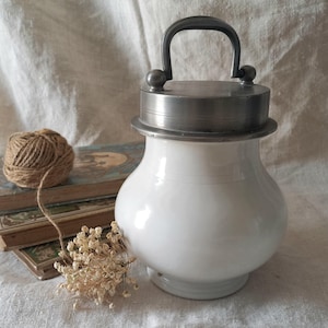 May include: A white ceramic jar with a metal lid and handle. The jar has a rounded body and a pedestal base. The lid is silver-colored. A small bundle of dried flowers and a ball of twine are next to the jar.