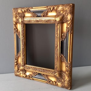 Op de afbeelding: Omlijste gouden fotolijst met een zwart interieur. De lijst heeft gedetailleerde, gebeeldhouwde versieringen en een rijke, antieke uitstraling. De lijst is gemaakt van een stevig materiaal en is ontworpen om een foto of kunstwerk in te plaatsen.