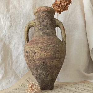 Peut inclure: Un vase amphore vieilli à deux anses, dans des tons de vert, de brun et de beige. Le vase antique présente une surface texturée et est orné d'une fleur séchée. Le vase repose sur une partition, évoquant une esthétique vintage.