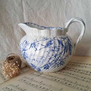 Pode incluir: Um jarro de cerâmica branca com um padrão floral azul de transferência e um corpo canelado. O recipiente tem uma pega curva, uma borda recortada e um bico. O design azul apresenta flores detalhadas e uma paisagem no exterior.