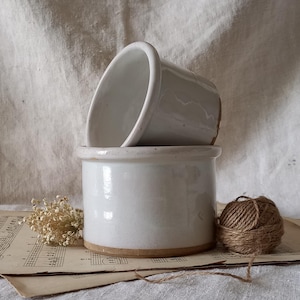 May include: Two white ceramic butter dishes, one stacked on top of the other. The bottom dish is cylindrical with a wide rim, and the top dish is a smaller, inverted cone shape. The dishes are on a sheet of vintage sheet music.
