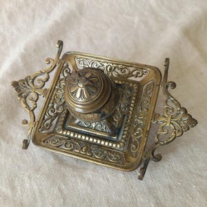 Pode incluir: Um tinteiro quadrado de latão com uma borda de filigrana ornamentada e pegas laterais decorativas. O recipiente central de tinta tem uma tampa abobadada com dobradiças e um motivo floral. A superfície metálica apresenta uma pátina.