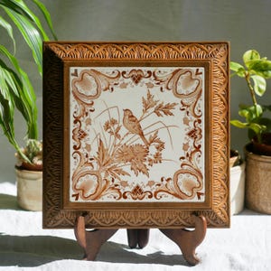 Può includere: Una cornice in legno marrone con una piastrella in ceramica bianca con un uccello marrone e un motivo floreale. La piastrella è su un supporto in legno.