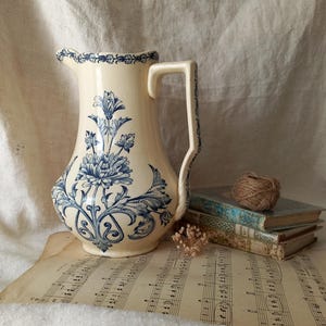 Pode incluir: Uma jarra de cerâmica na cor creme com um padrão floral azul, apresentando um grande motivo de crisântemo na parte frontal e uma borda decorativa ao longo do bocal. A jarra possui uma alça curva e um bico com detalhes botânicos em azul.