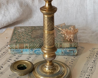 May include: A brass candlestick holder featuring a cylindrical stem with an intricate geometric pattern, a circular base with concentric ring detailing, and a separate matching circular drip tray resting beside the main candle holder base.