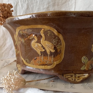 Op de afbeelding: Een bruine keramische plantenbak met een rechthoekige vorm en decoratieve voetjes. De plantenbak heeft een reliëfontwerp van twee vogels in een omlijst paneel. De rand en details zijn geaccentueerd met een lichtere kleur.