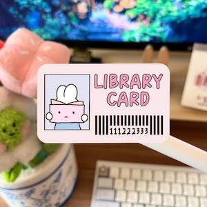 May include: A pink library card sticker with the words "LIBRARY CARD" in pink. The sticker features a cartoon bunny holding a book and a barcode. The background is blurred, with a keyboard and other objects visible.