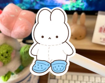 May include: A white bunny sticker with black outlines, wearing light blue cartoon clogs. The sticker has a minimalist design with stitched details. The bunny has small black eyes and a small black nose. The sticker is on a desk with other objects in the background.