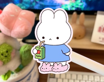 May include: A white bunny sticker wearing a blue shirt and pink bunny slippers, holding a green apple juice box with the word "aple" on it. The sticker has a white border and is on a desk with other items in the background.