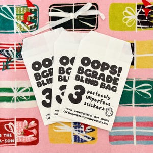 May include: Three white paper bags with the text "OOPS! BGRADE BLIND BAG" and "3 perfectly imperfect stickers" in black. The bags are on a pink background with gift box patterns.