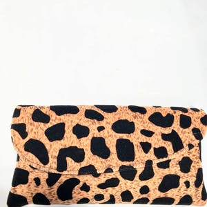 May include: A rectangular clutch bag with a flap closure. The bag has a tan background with black spots, resembling a leopard print. The bag is made of a soft material.