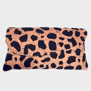 May include: A rectangular clutch bag with a fold-over flap. The bag has a tan background with irregular black spots, creating a leopard print design. The clutch is made of a soft, textured material.