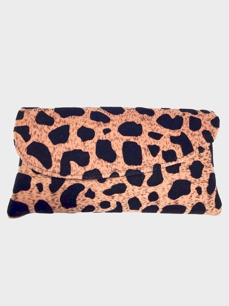 May include: A rectangular clutch bag with a fold-over flap. The bag has a tan background with irregular black spots, creating a leopard print design. The clutch is made of a soft, textured material.