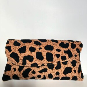 May include: A rectangular clutch bag with a flap closure. The bag has a tan background with large black spots, resembling a leopard print. The clutch is made of a soft material and is designed to be folded over.