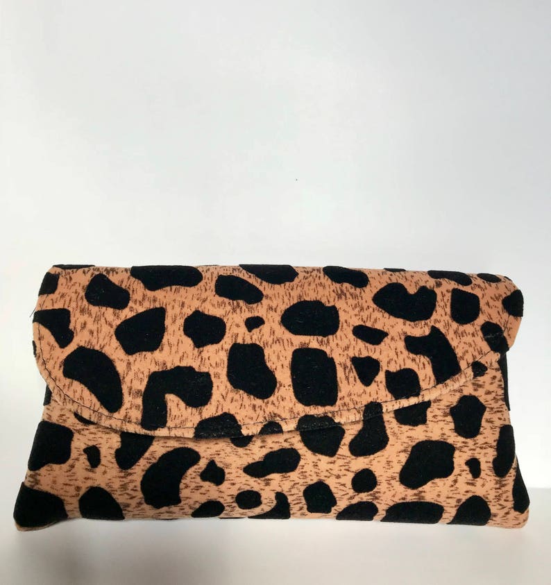May include: A rectangular clutch bag with a flap closure. The bag has a tan background with large black spots, resembling a leopard print. The clutch is made of a soft material and is designed to be folded over.