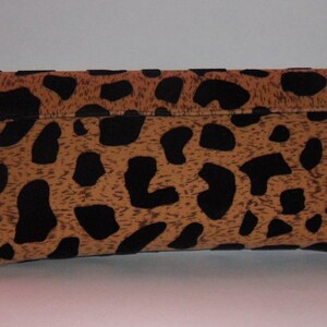May include: A rectangular clutch bag with a fold-over flap. The bag has a tan background with black leopard spots. The clutch is made of a textured material and is set against a white background.