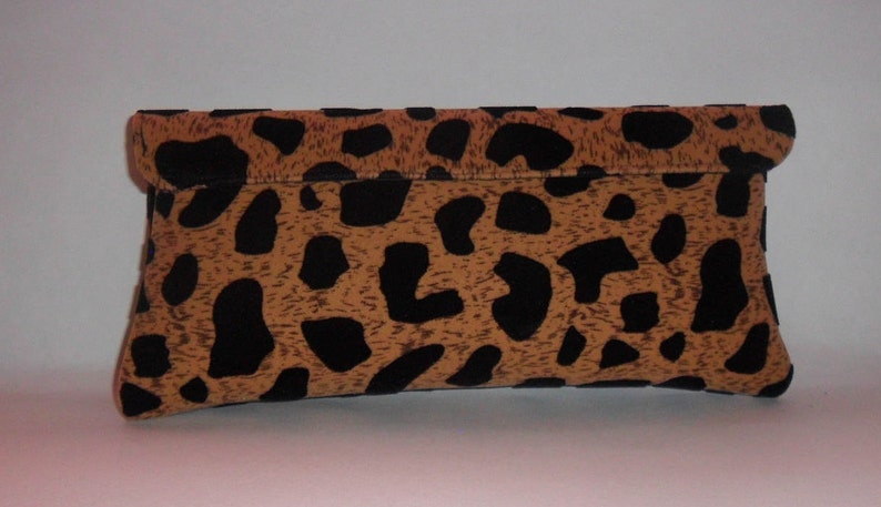 May include: A rectangular clutch bag with a fold-over flap. The bag has a tan background with black leopard spots. The clutch is made of a textured material and is set against a white background.