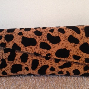 May include: A fold-over clutch bag featuring a tan and black animal print. The rectangular bag has rounded corners and a flap closure. The fabric displays a spotted pattern.