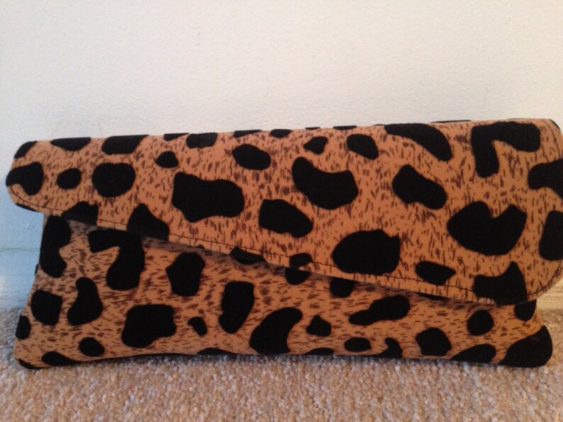 May include: A fold-over clutch bag featuring a tan and black animal print. The rectangular bag has rounded corners and a flap closure. The fabric displays a spotted pattern.