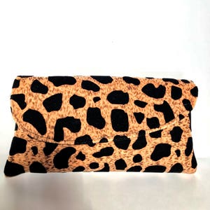 May include: A rectangular clutch bag with a flap closure. The bag has a tan background with black spots, reminiscent of a leopard print. The bag is made of a soft fabric.