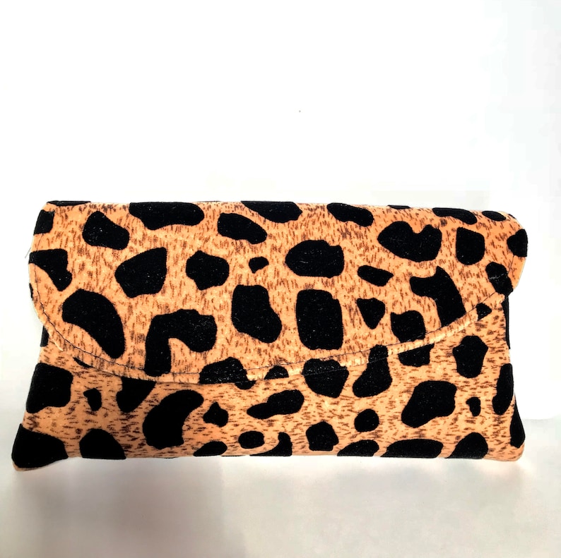 May include: A rectangular clutch bag with a flap closure. The bag has a tan background with black spots, reminiscent of a leopard print. The bag is made of a soft fabric.