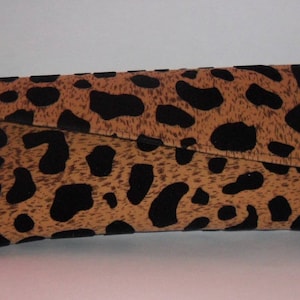 May include: A leopard print clutch bag. The bag is a rectangular shape with a folded-over flap. The pattern features black spots on a tan background. The bag is made of a soft fabric.