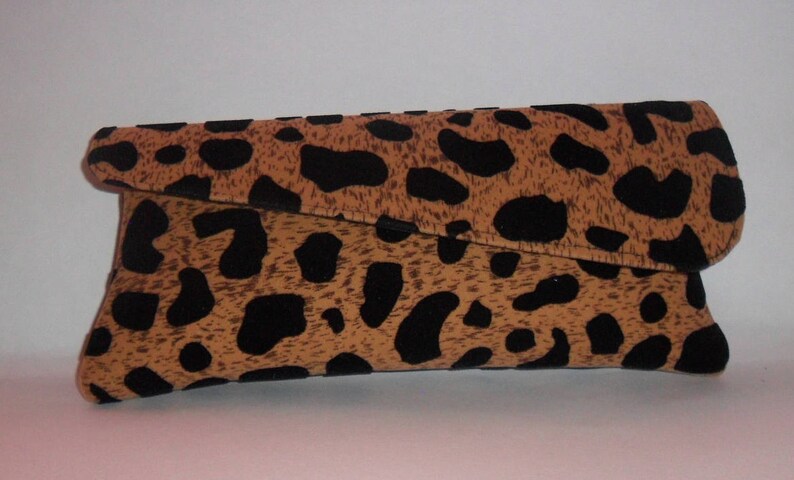 May include: A leopard print clutch bag. The bag is a rectangular shape with a folded-over flap. The pattern features black spots on a tan background. The bag is made of a soft fabric.