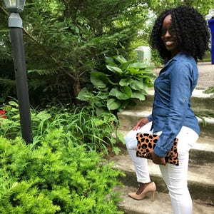 May include: A denim shirt and white trousers outfit. The look is completed with nude heels and a leopard print clutch. The person is standing on outdoor steps with greenery in the background.
