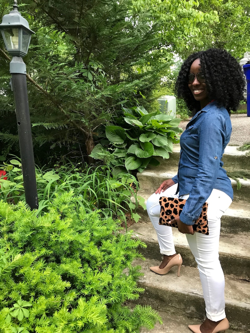 May include: A denim shirt and white trousers outfit. The look is completed with nude heels and a leopard print clutch. The person is standing on outdoor steps with greenery in the background.