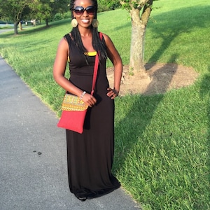 May include: A woman wearing a black maxi dress with a red crossbody bag. The bag has a colorful patterned flap. She is wearing sunglasses and large gold earrings. The background is a green grassy area with trees.