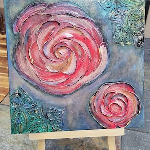 May include: An abstract painting on canvas featuring two roses in shades of red, pink, and gold. The textured artwork includes decorative teal accents and is displayed on a small wooden easel.