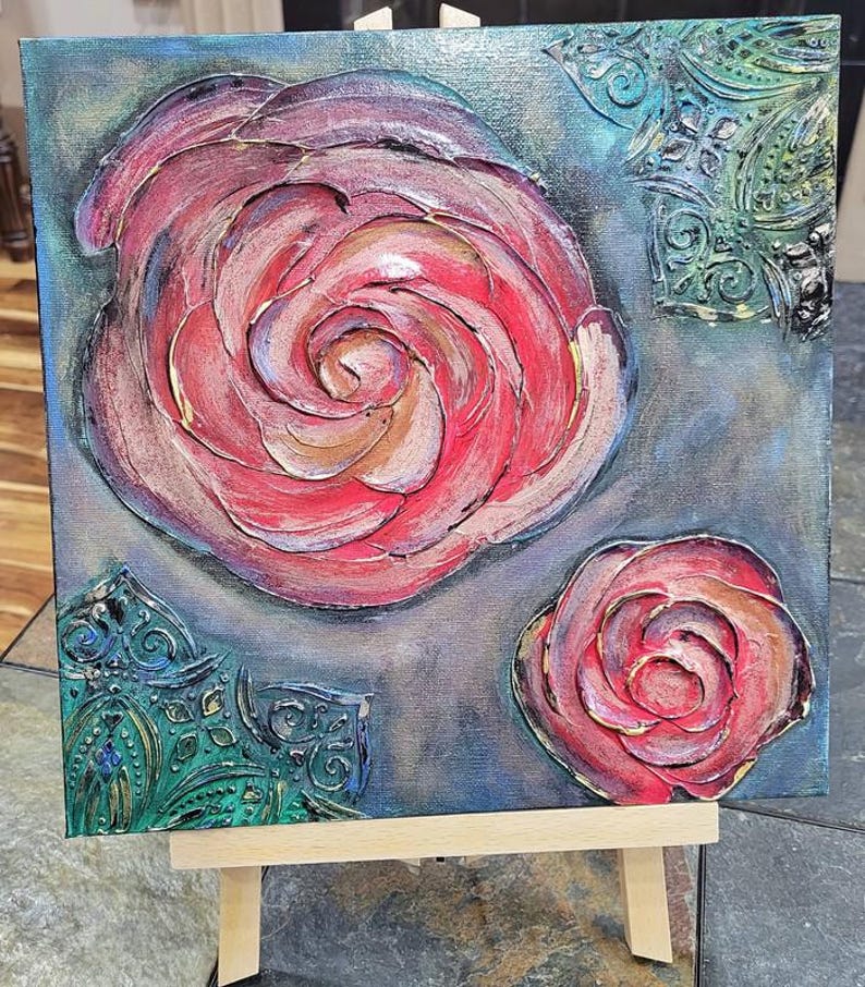 May include: An abstract painting on canvas featuring two roses in shades of red, pink, and gold. The textured artwork includes decorative teal accents and is displayed on a small wooden easel.