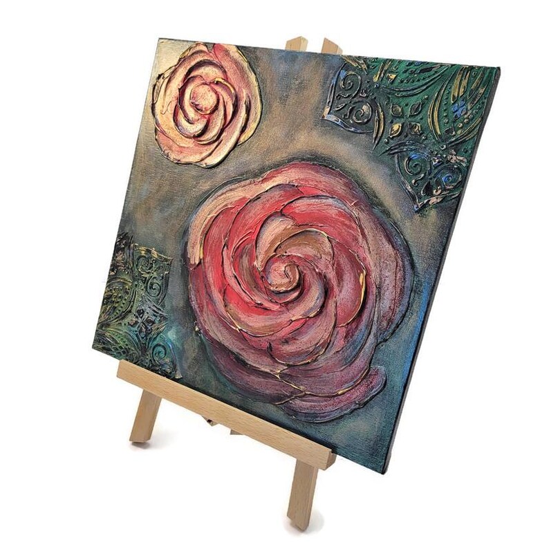 May include: Square canvas art featuring textured roses in shades of red, pink, and peach, set against a muted gray-blue background. Decorative green and gold corner accents add detail. The artwork is displayed on a small wooden easel.