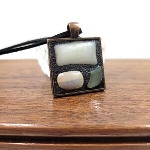 May include: A square pendant necklace with a black leather cord. The pendant is made of metal with a copper finish and features three different colored stones. The stones are white, iridescent, and green.