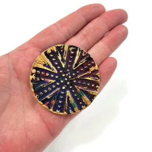May include: A round, gold-toned brooch with a textured surface. The brooch is decorated with a pattern of gold dots and lines, and has a dark blue, purple, and green background.