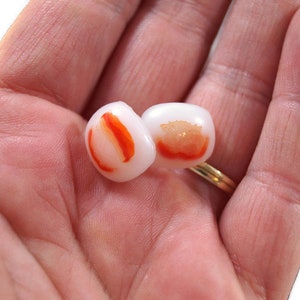 May include: Two white and orange glass beads with a red and orange swirl design. The beads are in the palm of a hand.