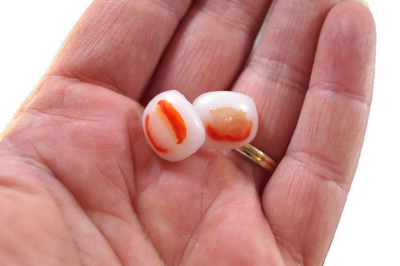 May include: Two white and orange glass beads with a red and orange swirl design. The beads are in the palm of a hand.