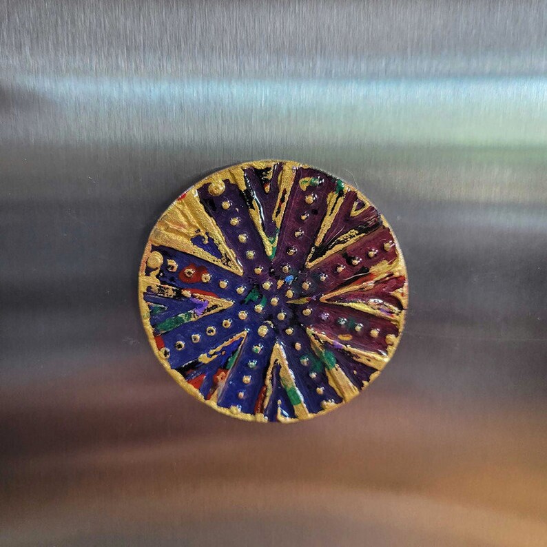 May include: A round, colorful, decorative magnet with a gold, purple, blue, green, and red design. The magnet has a textured surface and a circular shape.