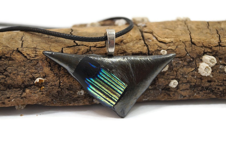 May include: A triangular pendant necklace with a black finish and a section of iridescent blue and green glass. The pendant is suspended from a black cord and has a silver-colored bail. The necklace is displayed on a piece of driftwood.