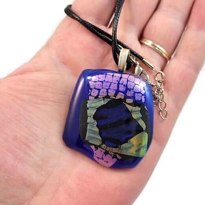 May include: A square glass pendant necklace with a blue, black, pink, and green mosaic design. The pendant is attached to a black cord.