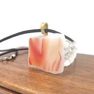 May include: A white and orange agate pendant necklace with a black cord and a gold-toned clasp. The pendant is a square shape with a marbled pattern.