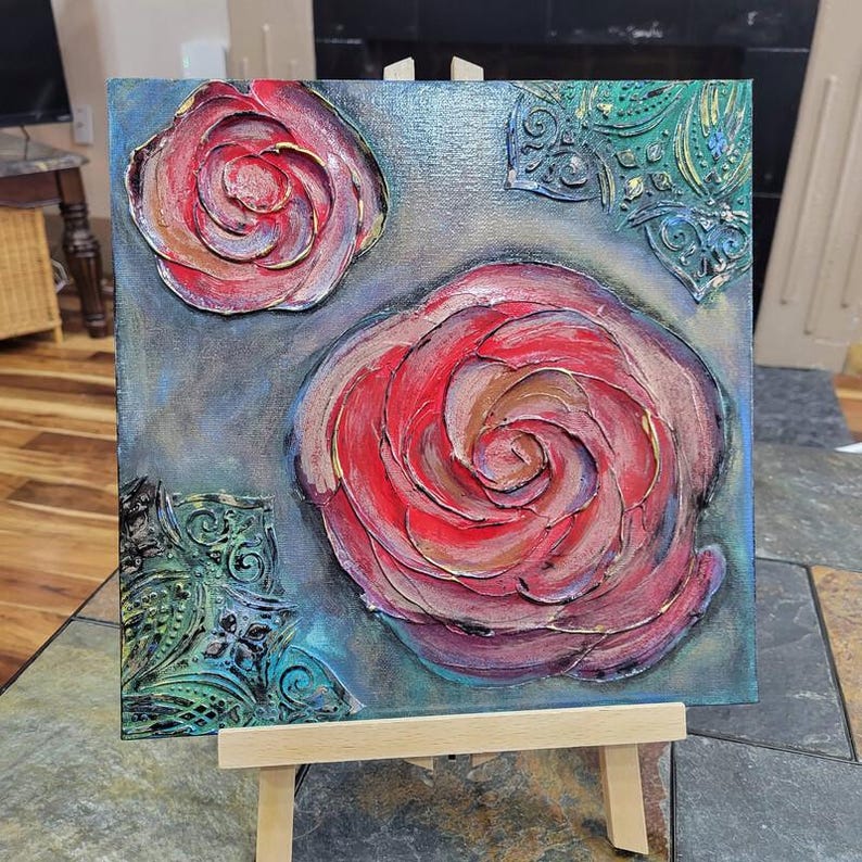 May include: Square canvas art featuring textured, raised roses in shades of red, pink, and gold. The background is a blend of blue and gray, with decorative green and gold accents in the corners. The artwork is displayed on a small wooden easel.