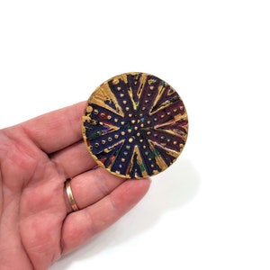 May include: A round, gold-colored button with a purple, blue, and green geometric design. The button has a textured surface and is decorated with small gold dots.