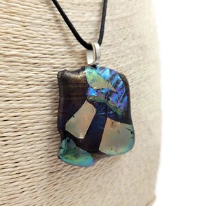 Puede incluir: Un collar con un colgante cuadrado de vidrio negro, iridiscente y dorado. El colgante está hecho de vidrio fundido y tiene una anilla de plata.