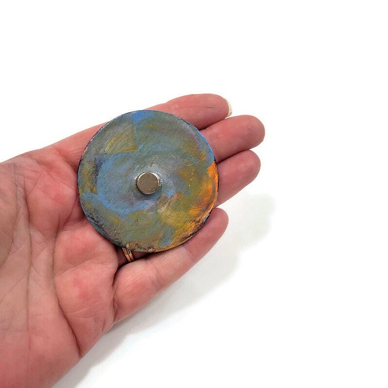 May include: A round, metallic disc with a blue, green, and orange paint design. A small, silver magnet is attached to the center of the disc.